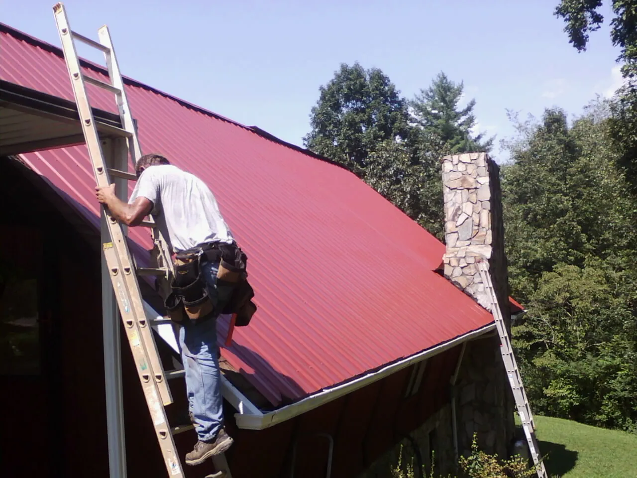 Roofer on a ladder beside a red metal roof and stone chimney — Soffit & Fascia Repair by Roofing Brothers in Stittville